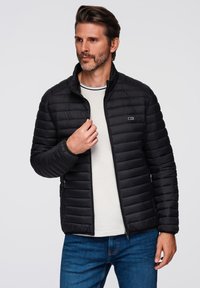Chaqueta negra acolchada con cuello alzado, cierre de cremallera en la parte delantera y bolsillos laterales. Lleva puesta una camisa de rayas en colores claros con unos pantalones de mezclilla.