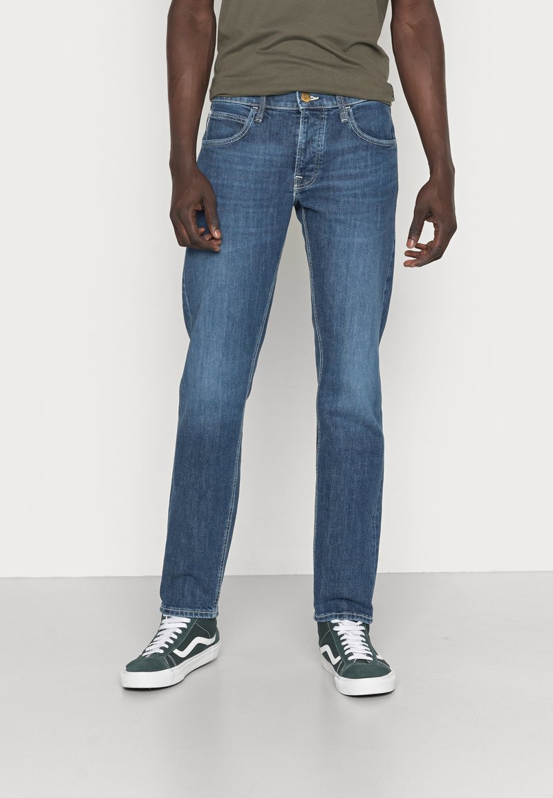 Blue denim jeans with a straight leg cut, featuring five pockets and golden hardware, paired with dark green sneakers.