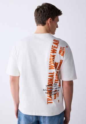 Hombre que lleva una camiseta blanca de manga corta con texto vertical en naranja y negro que dice "Traditional Workwear" en la espalda, combinado con vaqueros azules.