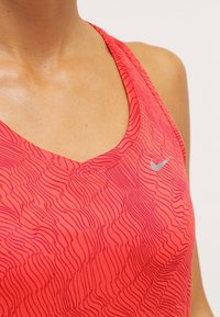 Débardeur Nike corail vif avec un motif ondulé, doté d'un décolleté en U et de fines bretelles. Agrémenté d'un petit logo argenté sur la poitrine.