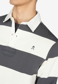 Polo a maniche lunghe con righe orizzontali blu navy e bianche. Presenta un colletto bianco, una serie di bottoni e un emblema ricamato sul petto.