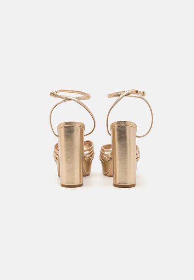 Sandalias de tacón alto metálicas doradas con un tacón grueso, diseño de tiras entrelazadas y correa de tobillo ajustable. Textura suave.