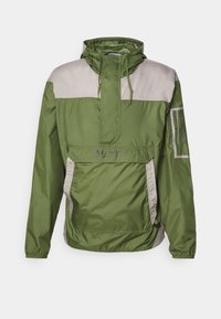Chaqueta cortaviento verde y gris claro con capucha, cierre parcial con cremallera, bolsillo frontal y bolsillo lateral de malla. Tejido suave y ligero.