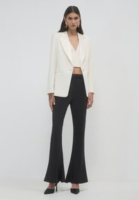 Frau in einem weißen Blazer und Crop-Top, mit schwarzen Schlaghosen und schwarzen High Heels, steht vor einem einfarbigen weißen Hintergrund.