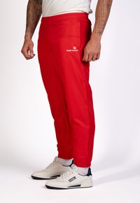 Rode sportieve broek met een elastische tailleband, smal gesneden pasvorm en ritsen bij de enkels. Heeft een wit logo op de linkerdij.