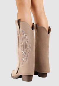 Bottes de cowboy en suède beige avec un design de plume stylisé en broderie blanche, présentant un bout arrondi et un talon bloc de hauteur intermédiaire.
