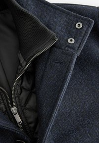 Marineblauer Wollmischmantel mit wattiertem schwarzem Innenfutter, Reißverschluss und geripptem Kragen. Druckknöpfe und strukturierte Stoffdetails vorhanden.