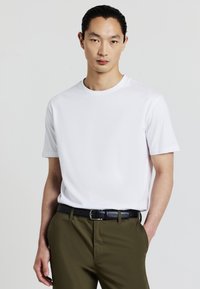Giovane uomo con capelli pettinati all'indietro che indossa una maglietta bianca a maniche corte, semplice, dentro dei pantaloni verde oliva con una cintura nera.