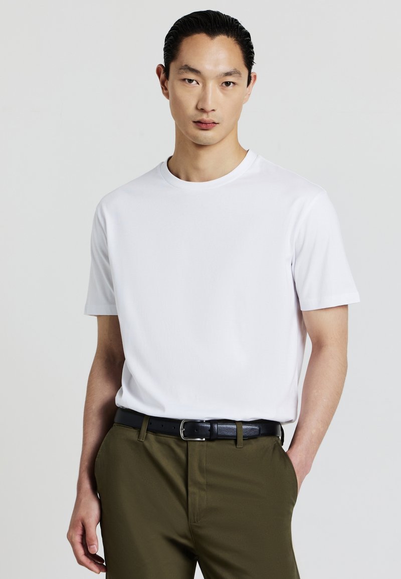 Giovane uomo con capelli pettinati all'indietro che indossa una maglietta bianca a maniche corte, semplice, dentro dei pantaloni verde oliva con una cintura nera.