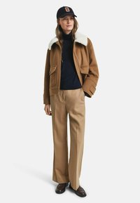 Giacca di lana marrone con colletto in shearling bianco, dolcevita blu, pantaloni beige a gamba larga e scarpe in pelle marrone. Cappellino blu con la lettera arancione G.