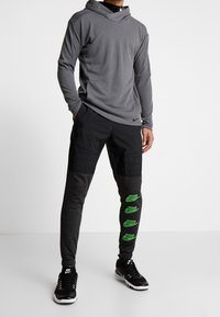 Graues langärmliges Kapuzenshirt kombiniert mit schwarzen und anthrazitfarbenen Hosen mit strukturierten Akzenten und grünen Logo-Designs, abgerundet mit schwarzen Sneakern.