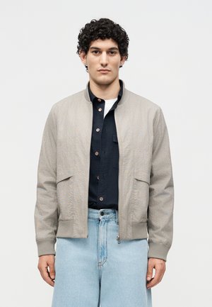 Jeune homme aux cheveux bouclés portant une veste bomber beige sur une chemise noire boutonnée et un jean taille haute bleu clair, debout devant un fond blanc.