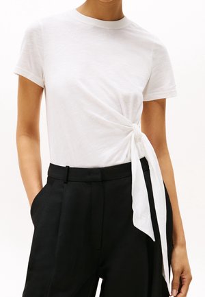 Weißes T-Shirt mit kurzen Ärmeln und Knotendetail an der Seite, kombiniert mit hoch taillierter schwarzer Hose, Hände in den Taschen.