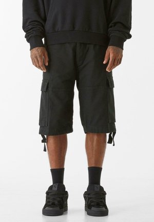 Shorts cargo noirs avec plusieurs poches, longueur genou, fabriqués dans un tissu durable, dotés de cordons de serrage réglables aux ourlets. Portés avec des baskets noires.