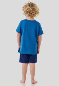 Ein kleines Kind mit lockigem blonden Haar steht barfuß, trägt ein blaues T-Shirt und marineblaue Shorts und schaut mit dem Rücken zu einer schlichten hellen Hintergrun.