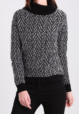 Femme portant un pull à col roulé à manches longues à motifs noir et blanc avec un pantalon noir, debout devant un fond uni.