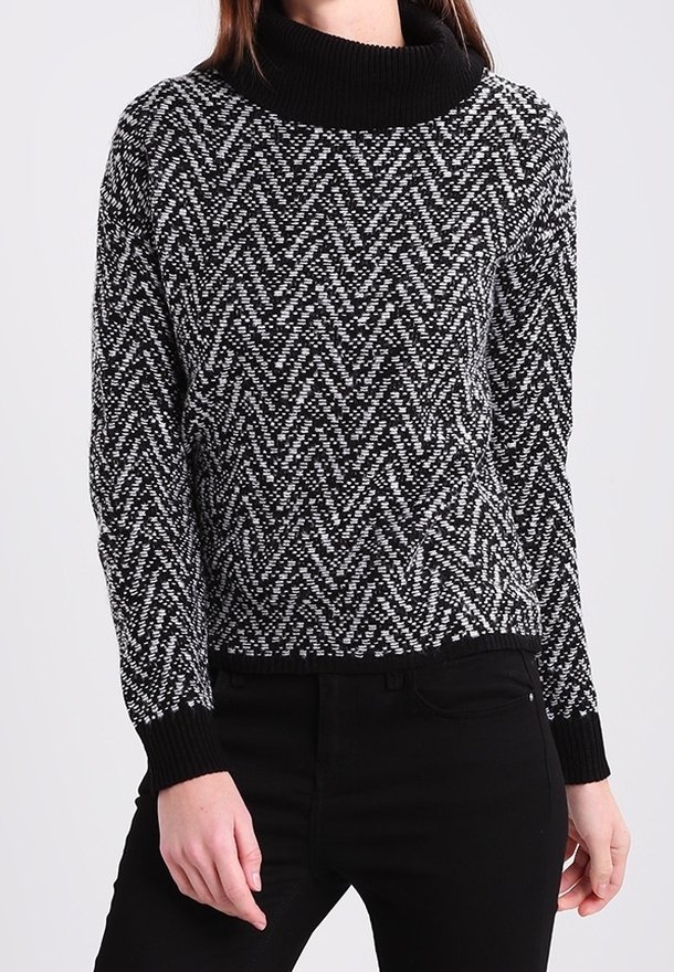 Femme portant un pull à col roulé à manches longues à motifs noir et blanc avec un pantalon noir, debout devant un fond uni.