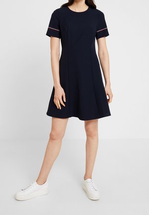 Marineblaues Kleid mit kurzen Ärmeln, tailliertem Oberteil und ausgestelltem A-Linien-Rock. Verziert mit kontrastierenden Nähten an den Ärmeln. Kombiniert mit weißen Turnschuhen.