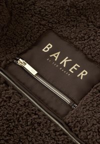 Un tessuto in pile marrone strutturato circonda un rettangolo marrone con la scritta "BAKER" in oro e una cerniera, con accessori dorati.