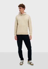 Sudadera beige con bolsillo canguro y logo en el pecho, combinada con pantalones negros y zapatillas multicolores. Diseño de corte recto.