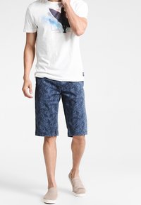 Vit t-shirt med surfgrafik, blå mönstrade shorts och ljusbruna slip-on skor. Shortsen har ett ormbunksmönster.
