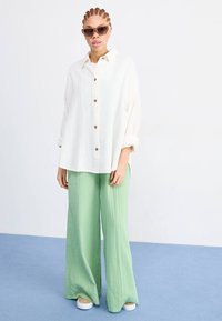 White crinkled button-up shirt with brown buttons, paired with light green wide-leg pants. Sunglasses complement the outfit.