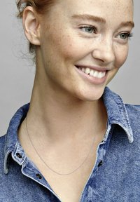 Strieborný reťazový náhrdelník na modeli so svetlou pokožkou a pehami, ktorý má na sebe džínovú košelu. Košeľa má špicatý golier a čierne gombíky.