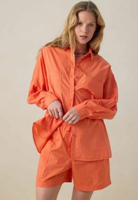 Chemise orange à manches longues, avec deux poches poitrine et boutonnière, accompagnée de shorts orange assortis avec poches latérales et taille élastique.