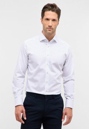 Camicia bianca con un piccolo motivo a pois viola, maniche lunghe, colletto a bottoni e taglio dritto; abbinata a pantaloni blu scuri.
