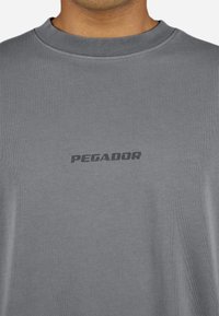 Tricou gri cu mânecă scurtă, realizat dintr-un material ribbed, având un logo subtil de culoare neagră "PEGADOR" pe zona pieptului și un guler rotund.