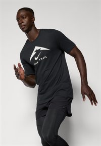 Schwarzes T-Shirt mit kurzen Ärmeln und einem weißen Logo, das ein dreieckiges Design zeigt, kombiniert mit schwarzen Sportshorts und enganliegenden Leggings.