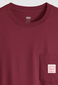 Μπλούζα Levis σε χαλαρή γραμμή σε μπορντό χρώμα, με στρογγυλή λαιμόκοψη και μπροστινή τσέπη που φέρει λευκή ετικέτα με κόκκινο κείμενο και γραφικό.