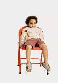 Barn sitter på en röd metallstol, iförd en beige sweatshirt med färgglada mönster och rosa shorts, tillsammans med ljusgrå sandaler.