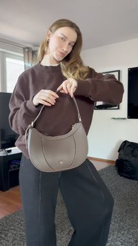 Jonge vrouw binnen met een grijze leren handtas met een enkele schouderriem, gekleed in een bruine trui en zwarte wijde broek.