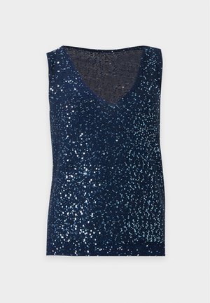 Femme aux longs cheveux foncés portant un haut sans manches à sequins bleu marine, une jupe midi en jean foncé et des chaussures Mary Jane noires, debout les mains dans les poches.