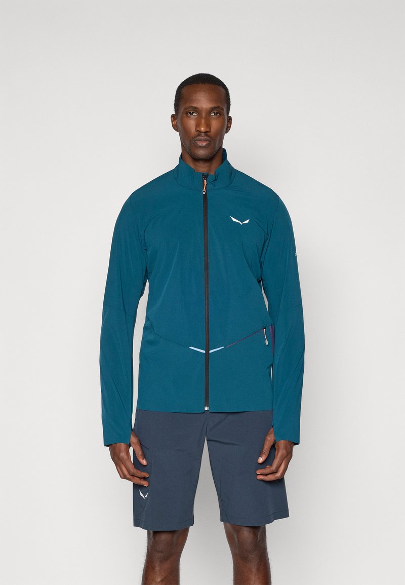 Τελείως γαλάζια ελαφριά ζακέτα με υψηλό γιακά και πλήρη φερμουάρ μπροστά, διαθέτοντας ανακλαστικές λεπτομέρειες, συνδυασμένη με σκούρα σορτς.