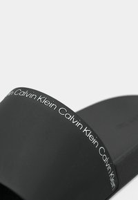 Fekete csúszásmentes szandál matt felülettel, széles pánttal, amelyet ismétlődő fehér "Calvin Klein" márkázás díszít. Simára textúrázott talp.