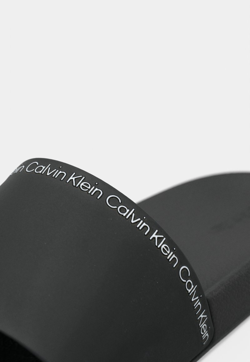 Sandale noire à talon plat avec une finition mate, présentant une large sangle ornée d'un logo "Calvin Klein" répété en blanc. Semelle intérieure lisse et texturée.