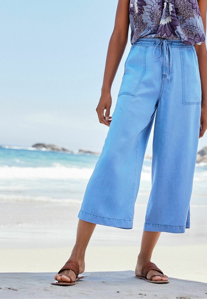 Helle blaue, weite Jeanshose mit Gummizug und großen Fronttaschen, kombiniert mit braunen, geflochtenen Sandalen in einer Strandkulisse.