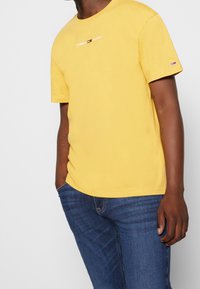 Yellow cotton t-shirt with short sleeves featuring "TOMMY JEANS" logo on chest. Worn with blue jeans. Slim fit and smooth texture.