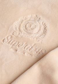 Tessuto beige chiaro con una trama morbida e liscia, caratterizzato da un logo e un disegno a corona in rilievo al centro, con la scritta "Superdry New York City".