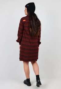 Robe à motif rouge et noir avec des manches longues, présentant des designs géométriques et un ourlet arrondi. Portée avec un bonnet noir et des chaussures.