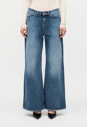 Személy széles szárú kék farmert, bézs színű hosszú ujjú felsőt és fekete hegyes orrú cipőt visel, sima háttér előtt áll.