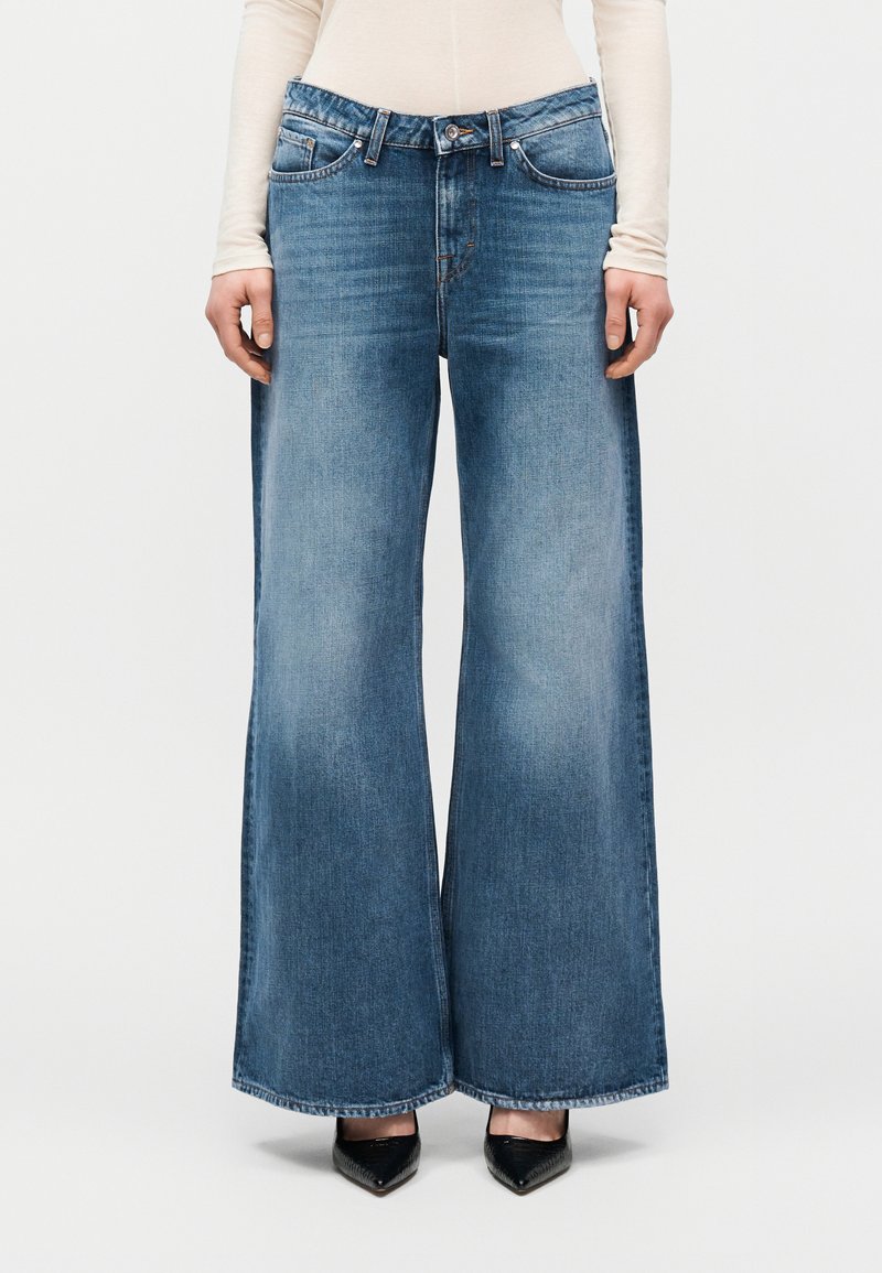 Άτομο που φοράει μπλε τζιν παντελόνι με φαρδιά μπατζάκια, μπεζ μακρυμάνικη μπλούζα και μαύρα παπούτσια με μυτερή μύτη, στέκεται μπροστά σε ένα απλό φόντο.