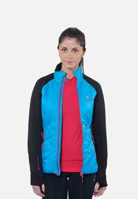 Femme portant une veste matelassée bleue avec des manches noires et une chemise rouge en dessous, debout devant un fond blanc uni.