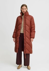 Langer, rostfarbener Daunenmantel mit Rautenmuster, hohem Kragen und Reißverschluss. Getragen über einem beigen Strickpullover und dunklen Hosen.