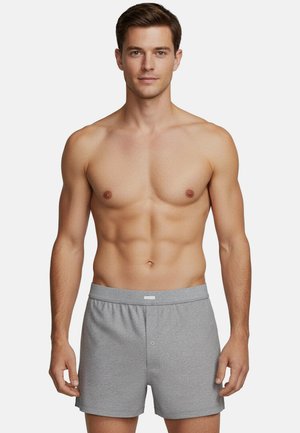 Grijze katoenen boxershorts met een elastische tailleband, voorzien van een knoopsluiting aan de voorkant en een gladde textuur. Model staat met een bloot bovenlichaam.