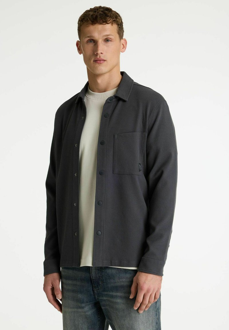 Graues, strukturiertes Hemd mit Kragen, vorne mit Knöpfen und einer einzelnen Brusttasche, über einem cremefarbenen T-Shirt getragen, kombiniert mit Jeans.