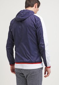 Námořnická modrá bunda s kapucí a bílými bočními panely, červenými akcenty a hladkým, lehkým materiálem; má stahovací kapuci a elastický spodní lem.