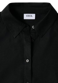 Chemise noire à manches longues avec col, fabriquée en tissu lisse. Présente des boutons argentés et une étiquette portant la mention "CECI". Design et texture minimalistes.